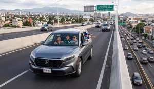 Una familia de cuatro personas en una SUV Honda plateada conduce por el segundo piso del Periférico Sur en CDMX, con vista de la ciudad y montañas.