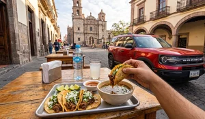 Tacos de birria recién servidos con consomé, cebolla, cilantro y limón; arquitectura tradicional tapatía al fondo, luz de mañana