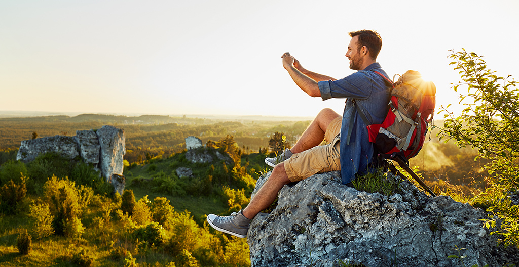 Hombre haciendo senderismo tomando foto con teléfono móvil sentado encima de la roca