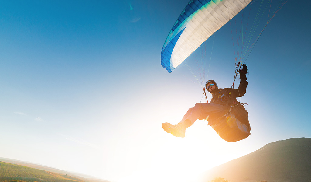 Un parapente despegará de una montaña con un toldo azul y blanco y el sol detrás.