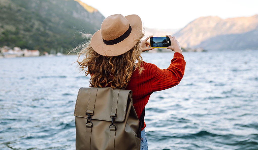 Mujer viajero en suéter casual que toma una foto de paisaje increíble con las montañas, el océano y el cielo de la puesta del sol en el smartphone