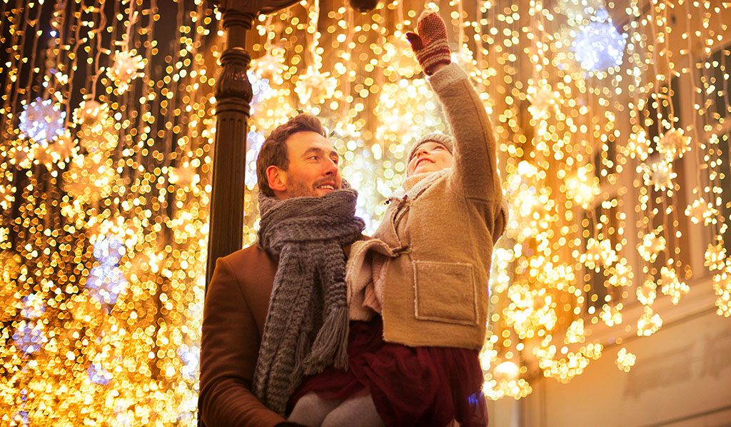 una sonriente en los brazos de su padre, con un abrigo marrón, pone las manos en la luz de la calle decorada