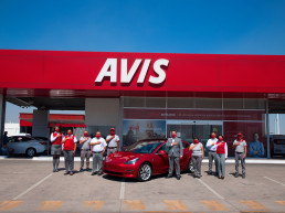 Plano general de trabajadores de Avis México rodeando un Tesla rojo Model 3 frente a una sucursal