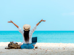 Imagen lifestyle mujer de espaldas con los brazos estirados al cielo sobre arena blanca en playa, vista al mar, haciendo freelance en su laptop, trabajando y relajándose, haciendo bleisure