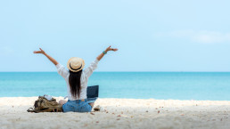 Imagen lifestyle mujer de espaldas con los brazos estirados al cielo sobre arena blanca en playa, vista al mar, haciendo freelance en su laptop, trabajando y relajándose, haciendo bleisure