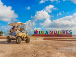 Pareja en carrito de golf frente a letrero de Isla Mujeres