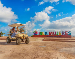 Pareja en carrito de golf frente a letrero de Isla Mujeres