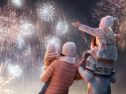 Fiesta de Año Nuevo. Familias felices, padres e hijas niñas están viendo fuegos artificiales. El niño se sienta en los hombros de su padre en un paseo nevado en la naturaleza. Vacaciones en invierno.