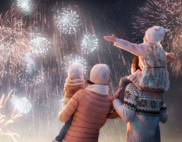 Fiesta de Año Nuevo. Familias felices, padres e hijas niñas están viendo fuegos artificiales. El niño se sienta en los hombros de su padre en un paseo nevado en la naturaleza. Vacaciones en invierno.